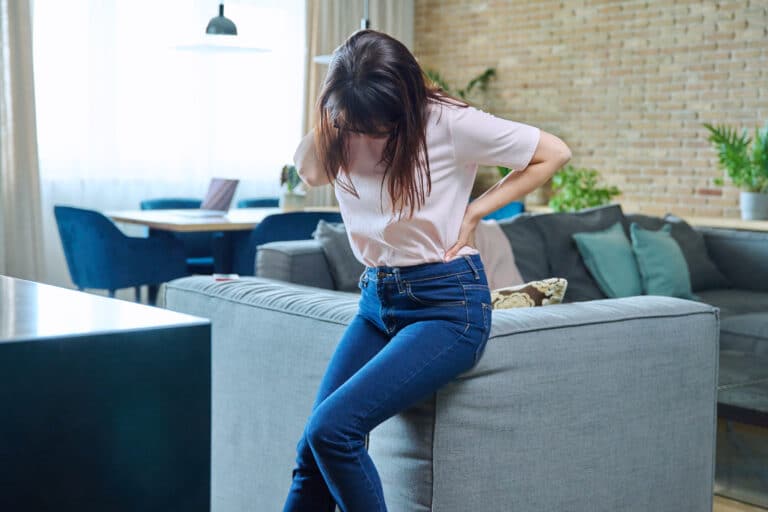 Woman in jeans and a light shirt holding her lower back in pain while sitting on a gray sofa, possibly experiencing discomfort from adult scoliosis or wondering if she is too old for a scoliosis brace.
