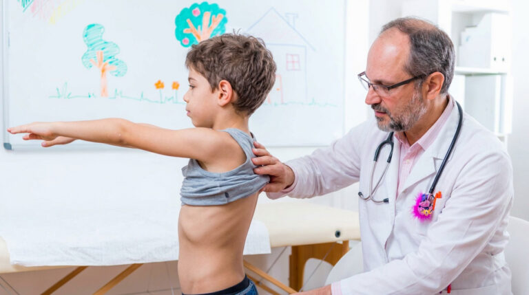 A doctor examines a young boy's back as he raises his arms in a medical office, highlighting the importance of scoliosis screening for early detection.