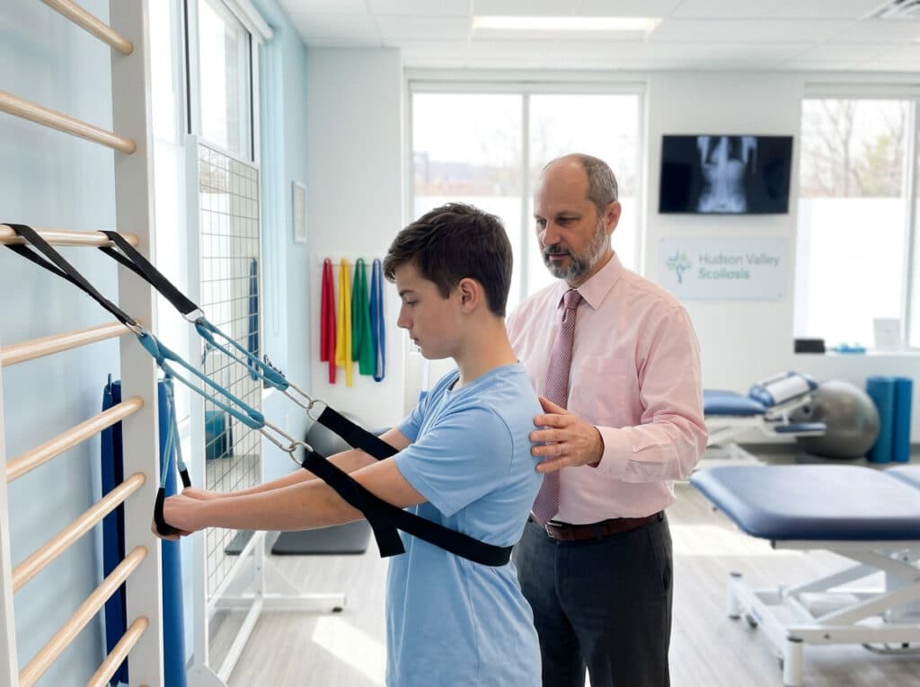 A boy does physical therapy exercises with straps as a therapist supervises in a clinic.